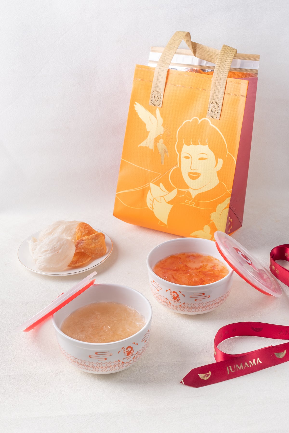 A gift set featuring two white bowls of soup or dessert, a plate with a pastry, a bright orange illustrated tote bag, and red ribbons on a clean white background.